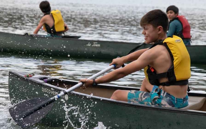 in the foreground, a boy paddles a canoe while two other boys paddle a canoe in the background